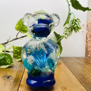 Peut inclure: Une figurine d'ours en résine bleue avec un corps transparent rempli de fleurs et de feuilles séchées. L'ours est debout sur une surface en bois.