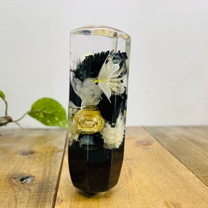 Peut inclure: Un cylindre en verre transparent rempli de fleurs séchées et de verdure. Les fleurs sont blanches et jaunes, et la verdure est vert foncé. Le cylindre est posé sur une surface en bois.