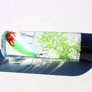 Pu&ograve; includere: Un cilindro trasparente con un tappo nero contiene un'esca da pesca verde e rossa e materiale vegetale verde.