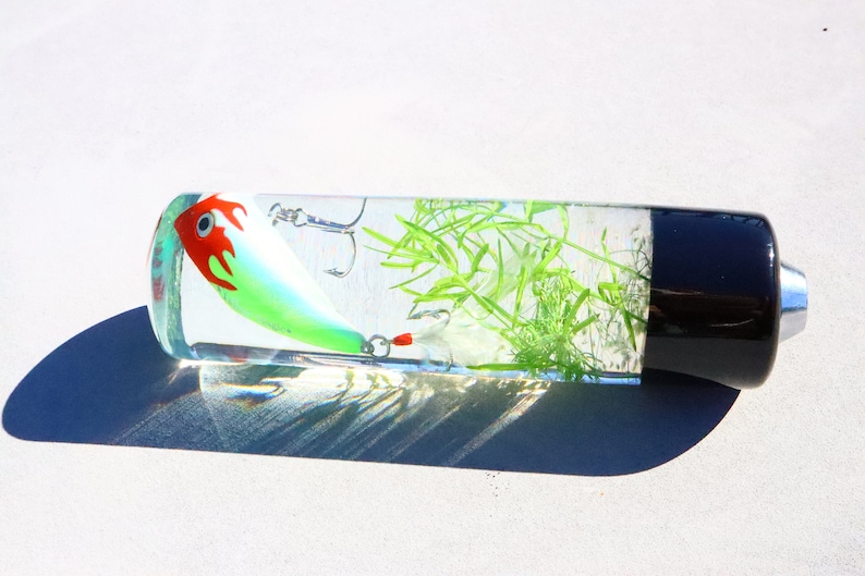 Pu&ograve; includere: Un cilindro trasparente con un tappo nero contiene un'esca da pesca verde e rossa e materiale vegetale verde.