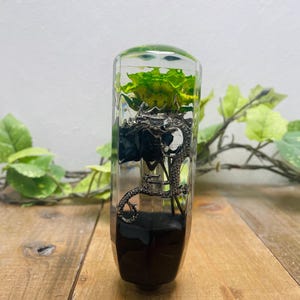 May include: A clear, rectangular resin art piece with a green and yellow flower and a silver dragon figurine. The base is dark brown. The piece is displayed on a wooden surface with green foliage in the background.