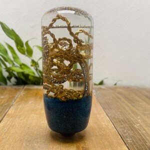 Puede incluir: Objeto cilíndrico transparente con cadenas doradas en su interior. La base es de color azul oscuro con purpurina. Se encuentra sobre una superficie de madera, con follaje verde al fondo. Mide unos 10 cm de alto.