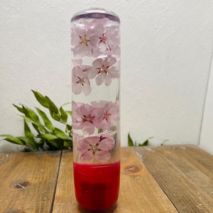 Puede incluir: Un pomo de palanca de cambios cilíndrico transparente con flores rosas en su interior. La parte inferior es de color rojo sólido. El pomo se muestra sobre una superficie de madera con follaje verde en el fondo.