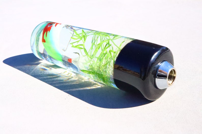 Pu&ograve; includere: Un pomello del cambio in vetro trasparente con un pesce rosso e verde all'interno e piante verdi. Il pomello ha una parte superiore nera con un'estremit&agrave; filettata cromata.