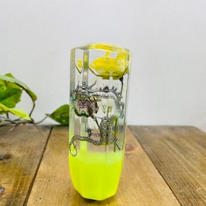 May include: A clear, rectangular resin object featuring a silver dragon sculpture and yellow floral elements. The bottom half is filled with bright neon yellow liquid. The object is displayed on a wooden surface.