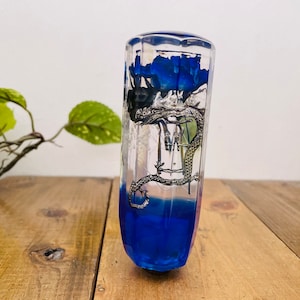 Op de afbeelding: Een transparante glazen versnellingspookknop met een blauwe harsbasis en een zilveren drakenontwerp. De draak houdt een blauwe bloem vast.