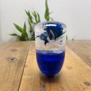 Puede incluir: Objeto de resina transparente de forma ovalada, con una base azul oscuro y una flor azul encapsulada. La parte superior es transparente, mostrando la flor y detalles blancos. El objeto está sobre una superficie de madera.