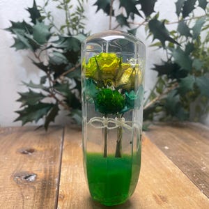 Könnte beinhalten: Ein durchsichtiger Glaszylinder mit grünem Sockel, gefüllt mit konservierten gelben und grünen Blumen. Die Blumen sind zu einem Strauß angeordnet und schweben in der Flüssigkeit.