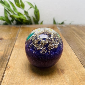 Puede incluir: Objeto esférico decorativo con una base de purpurina morada oscura. La esfera está adornada con cadenas doradas, pequeñas flores blancas y toques de verde y azul. Se encuentra sobre una superficie de madera.