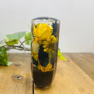 Op de afbeelding: Een transparante harscilinder met een zwarte basis gevuld met geperste gele en witte bloemen. De cilinder is ongeveer 15 cm hoog.