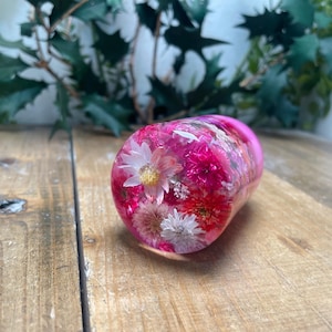 Puede incluir: Un tapón de botella de resina rosa con una variedad de flores prensadas rosas y blancas en su interior.