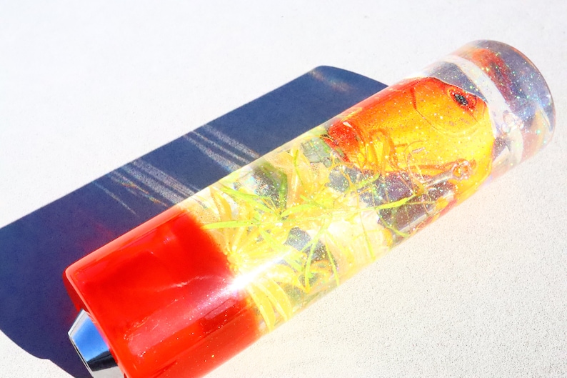 May include: A clear glass cylinder with a red base and a small metal cap. Inside the cylinder is a small orange fish, green plants, and glitter.
