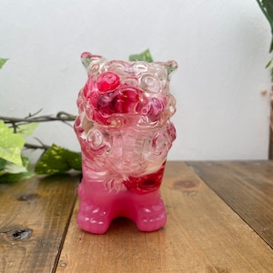Puede incluir: Una escultura de resina rosa y transparente de una criatura mítica con acentos florales. La escultura está detallada con diseños intrincados y presenta una superficie texturizada.