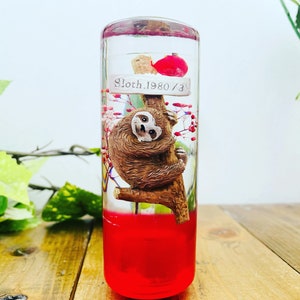 May include: A clear glass cylinder filled with a red liquid and a small sloth figurine hanging from a branch. The sloth is brown and has a white face. The cylinder has a white label with the text "Sloth.1980/3".
