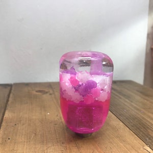 Peut inclure: Un pommeau de levier de vitesse en résine rose et violette avec un dessus transparent rempli de paillettes roses et violettes.