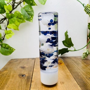 May include: A clear glass cylinder filled with white and blue flowers, preserved in resin. The cylinder has a white base and is sitting on a wooden surface.
