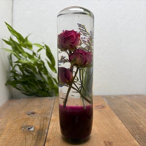 Peut inclure: Un presse-papier cylindrique en résine transparente contenant trois roses rouges séchées et de petites tiges de verdure. La base du cylindre présente une section opaque d'un magenta profond, tandis que la partie supérieure reste transparente, mettant en valeur la composition florale préservée.