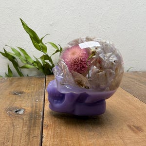 May include: A decorative skull sculpture with a clear resin dome filled with dried flowers. The base of the skull is a solid lavender color. Green leaves are in the background. The sculpture is on a wooden surface.