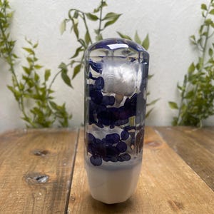 Könnte beinhalten: Ein klares, zylindrisches Objekt mit weißem Sockel, gefüllt mit dunkelblauen und weißen dekorativen Elementen. Die Oberseite ist abgerundet und das Objekt steht auf einer Holzoberfläche mit Grün im Hintergrund.