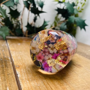 May include: A clear glass sphere filled with dried flowers and gold leaf. The flowers are pink, white, and purple. The sphere is sitting on a wooden surface.
