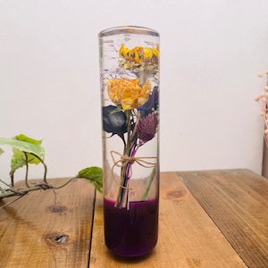 May include: A clear glass cylinder vase filled with dried flowers. The flowers are yellow, purple, and white. The vase has a purple base.