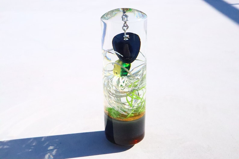 May include: A clear glass cylinder filled with a black guitar pick, green and white fishing line, and a dark brown liquid at the bottom.
