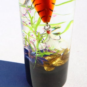 Może przedstawiać: Szklany słoik z przezroczystego szkła z przynętą wędkarską w pasy tygrysie, białymi pi&oacute;rami i złotymi kamieniami na dnie. Słoik jest wypełniony wodą i zielonymi i fioletowymi roślinami.