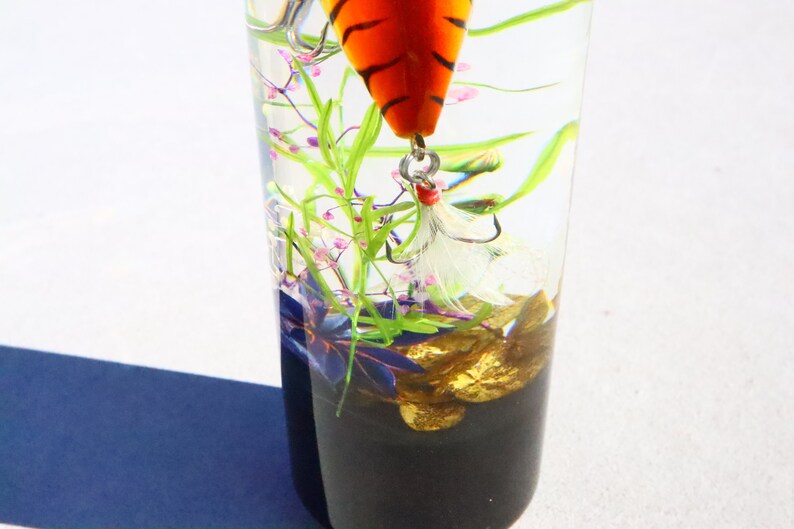 Może przedstawiać: Szklany słoik z przezroczystego szkła z przynętą wędkarską w pasy tygrysie, białymi pi&oacute;rami i złotymi kamieniami na dnie. Słoik jest wypełniony wodą i zielonymi i fioletowymi roślinami.