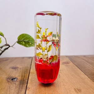 Op de afbeelding: Een transparante glazen pookknop met een rode harsbasis en geperste gele en groene bloemen erin.