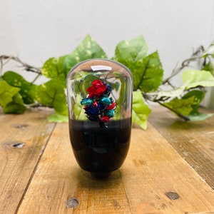 Può includere: Una sfera di vetro trasparente con una base nera, che contiene un arrangiamento floreale rosso, blu e verde. La sfera è su una superficie di legno.