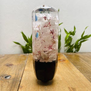 Könnte beinhalten: Ein klares, rechteckiges Objekt aus Harz mit dunkler Basis, gefüllt mit weißen und rosa Blütenblättern. Das Objekt wird auf einer Holzoberfläche mit grünem Laub im Hintergrund präsentiert. Die Oberseite ist klar.