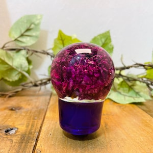 May include: A clear glass sphere filled with dried purple flowers, with a solid purple base. The sphere is sitting on a wooden surface.