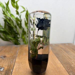 Op de afbeelding: Een heldere, rechthoekige hars papeterie met een zwarte roos en groen blad erin. De basis van de papeterie heeft een donkere, facetvorm. De achtergrond bevat groene bladeren en een houten oppervlak.