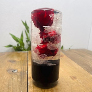 Puede incluir: Objeto decorativo cilíndrico transparente con base negra, que contiene flores rojas y delicados acentos blancos. La parte superior es redondeada y transparente, mostrando el arreglo floral. El objeto se exhibe sobre una superficie de madera.