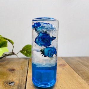 May include: A clear glass cylinder with a blue base filled with preserved blue roses and white lace. The cylinder is a decorative piece.