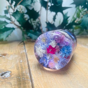 Könnte beinhalten: Ein klarer, ovaler Papierbeschwerer aus Harz, gefüllt mit einer Auswahl an bunten Trockenblumen. Die Blumen umfassen Farbtöne von Rosa, Blau und Weiß. Der Papierbeschwerer liegt auf einer Holzoberfläche, mit grünem Laub im Hintergrund.