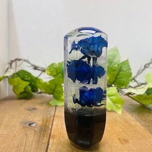 Pode incluir: Uma alavanca de câmbio de resina transparente com flores azuis encapsuladas dentro. A alavanca tem uma base preta.