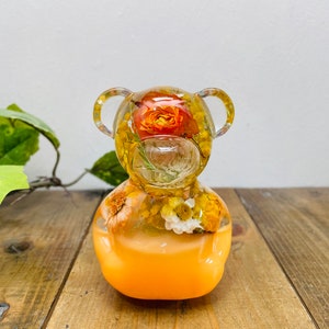Può includere: Una statuetta di orso in resina trasparente con un arrangiamento di fiori gialli e arancioni all'interno. L'orso è seduto su una superficie di legno.