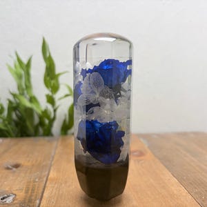 Puede incluir: Un pomo de palanca de cambios cilíndrico hecho de resina transparente, con flores de color azul intenso y delicadas hojas blancas esqueletizadas incrustadas en el material, con una base oscura y sólida en la parte inferior.