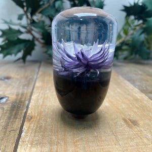 Può includere: Un pomello del cambio in vetro trasparente con un fiore viola all'interno. Il fiore è racchiuso in resina e ha un centro bianco.