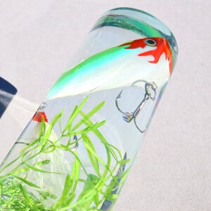 Pu&ograve; includere: Un'esca da pesca verde, bianca e rossa con un amo triplo &egrave; sospesa in un cilindro di vetro trasparente pieno d'acqua e piante di plastica verdi.