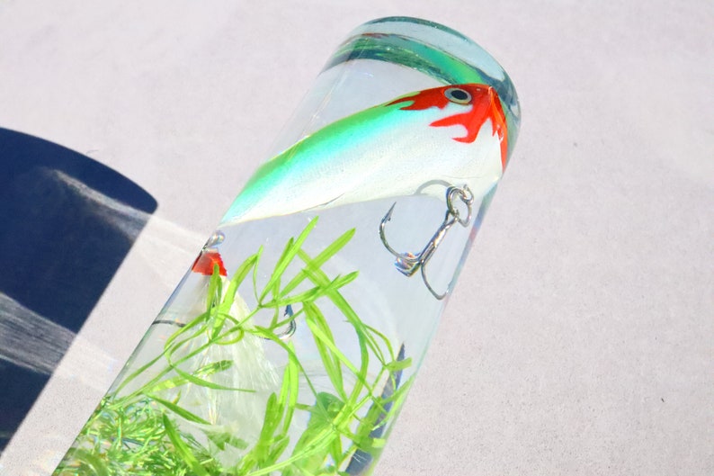 Pu&ograve; includere: Un'esca da pesca verde, bianca e rossa con un amo triplo &egrave; sospesa in un cilindro di vetro trasparente pieno d'acqua e piante di plastica verdi.