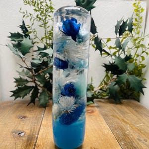 Könnte beinhalten: Ein durchsichtiger Glaszylinder gefüllt mit blauen und weißen Blumen, die in Harz konserviert sind. Der Zylinder hat eine blaue Basis und steht auf einer Holzoberfläche.