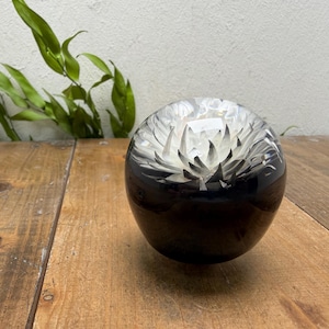 May include: A clear, spherical paperweight with a black base and a white, flower-like design inside. The paperweight sits on a wooden surface, with green leaves in the background.