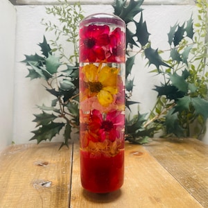 Op de afbeelding: Een transparante cilinder gevuld met geperste bloemen in tinten rood, geel en roze. De bloemen zijn verticaal in de cilinder gerangschikt, waardoor een kleurrijk en levendig display ontstaat.