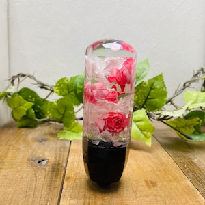 Op de afbeelding: Een transparante glazen versnellingspookknop met roze en witte bloemen ingebed in hars. De knop heeft een zwarte basis.