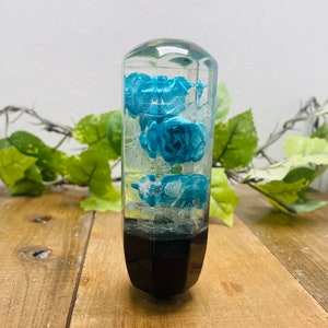 Op de afbeelding: Een transparante resin versnellingspookknop met blauwe bloemen erin. De knop heeft een zwarte basis.