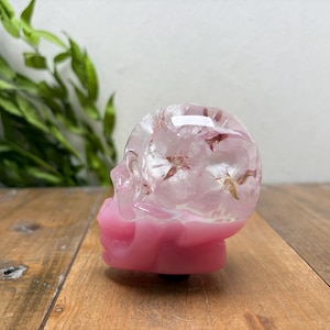 May include: A skull-shaped object made of clear resin with embedded pink flower petals in the upper portion and a solid opaque pink base. The object features a smooth, glossy finish and a distinct anatomical skull structure with visible eye sockets.