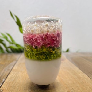 May include: A clear, cylindrical gear stick knob with a white base and a floral design. The floral arrangement features layers of white, pink, and green flowers encased in resin. The knob sits on a wooden surface.