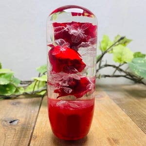 Peut inclure: Un cylindre en verre transparent rempli de roses rouges et de pétales blancs. Le cylindre est scellé en haut et en bas, et les roses sont conservées dans un liquide clair.